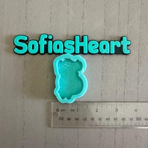 Puede incluir: Un molde de silicona azul claro con la forma de un personaje de dibujos animados, con una regla que muestra centímetros. El texto "SofiasHeart" está encima del molde en letras turquesas.