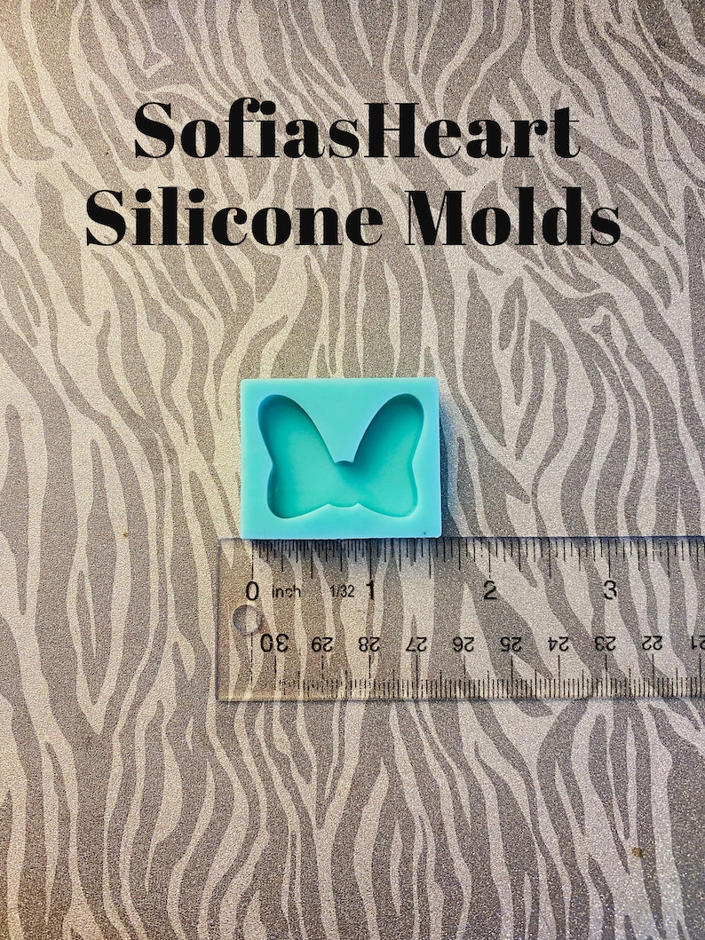 Puede incluir: Un molde de silicona azul claro con forma de lazo. El molde mide aproximadamente 2,5 cm de ancho y est&aacute; sobre una regla.