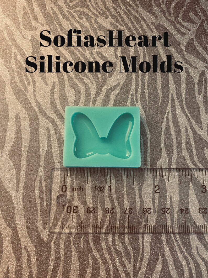 Puede incluir: Un molde de silicona azul claro con forma de lazo. El molde mide aproximadamente 1 pulgada de ancho y se muestra junto a una regla.