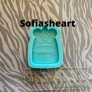 May include: A turquoise blue silicone mold of a small backpack with a bow on top. The mold is approximately 2 inches wide.