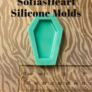 May include: A teal blue silicone mould shaped like a coffin. The mould is approximately 2.5 cm wide and 5 cm long. The mould is sitting on a brown and white patterned surface.