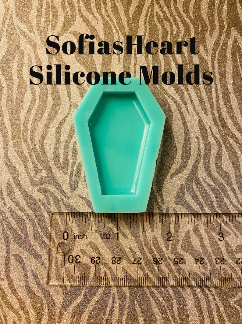 May include: A teal blue silicone mould shaped like a coffin. The mould is approximately 2.5 cm wide and 5 cm long. The mould is sitting on a brown and white patterned surface.