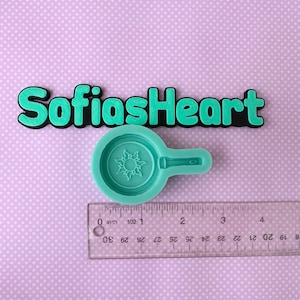 May include: A green silicone mold shaped like a frying pan with a sun design inside. The mold is on a purple background with a ruler for size reference. The text "SofiasHeart" is above the mold.