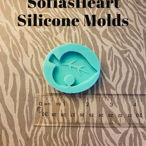 Puede incluir: Un molde de silicona azul claro con forma de hoja verde con un pequeño tallo marrón. El molde tiene un diámetro de aproximadamente 5 cm. El molde está sobre una superficie con estampado de cebra con una regla para la escala.
