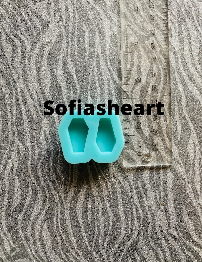 May include: A blue silicone mold with two coffin-shaped cavities. The mold is approximately 2.5 inches wide and 1.5 inches tall. The mold is on a gray and white patterned surface. The text "Sofiasheart" is visible in the image.