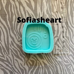 May include: A blue silicone mold shaped like an instant camera with a Mickey Mouse head in the viewfinder. The mold is approximately 2.5 inches tall.