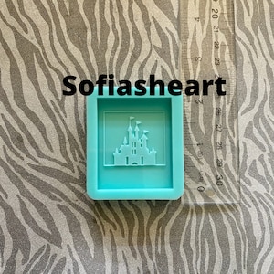 May include: A light blue silicone mold with a raised design of a castle with flags. The mold is approximately 2 inches wide and 2 inches tall.