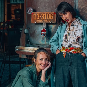 Op de afbeelding: Twee vrouwen staan voor een oldtimer. De vrouw links draagt een groene jas en heeft haar hand op haar kin. De vrouw rechts draagt een blauwe jas over een geprint shirt en jeans. De auto heeft een kentekenplaat met de tekst "13-3106 IOWA 1939".
