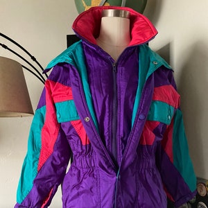 May include: A vintage, color-blocked jacket in purple, teal, pink, and magenta. The jacket features a high collar, a zipper closure, and snap details. The waist is cinched with an elastic band. The jacket is displayed on a mannequin.