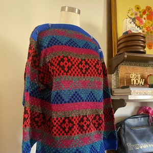 Könnte beinhalten: Ein langärmeliger Strickpullover mit einem lebendigen geometrischen Muster in Blau, Rot, Schwarz und Magenta. Der Pullover hat einen einfarbigen, gerippten blauen Halsausschnitt und Bündchen sowie eine Reihe kleiner blauer Knöpfe an der rechten Schulter.
