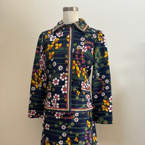 May include: A dark blue two-piece set consisting of a long-sleeve jacket and a skirt. The fabric features a repeating pattern of white, yellow, and pink flowers with green stems and thin horizontal lines. The jacket has gold-tone buttons.