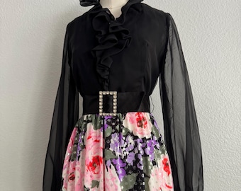 May include: A vintage black dress with a floral skirt. The dress has a sheer long-sleeved top with a ruffled collar and a black belt with a square rhinestone buckle. The skirt features pink, red, and purple flowers.