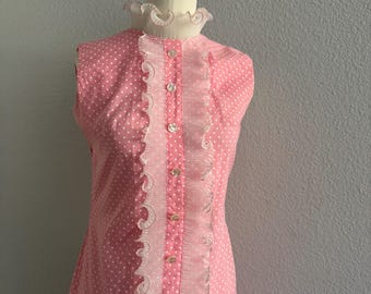 May include: A sleeveless pink dress featuring a white polka dot pattern, a high ruffled collar, and a central vertical placket adorned with light-colored buttons and decorative scalloped white trim running down the front of the garment.