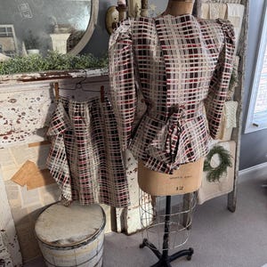 May include: A vintage-style two-piece outfit displayed on a mannequin. The set features a beige, red, and black plaid pattern. The top has long sleeves and a peplum, while the matching shorts are visible. The mannequin is on a metal stand.