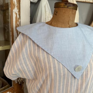 Könnte beinhalten: Ein kurzärmeliges Kleid mit einem einfarbigen, hellblauen Matrosenkragen, der an der Spitze mit einem einzelnen weißen Knopf versehen ist. Der Hauptteil des Kleides besteht aus einem Stoff mit vertikalen Streifen in hellblau und cremefarben.
