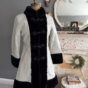 May include: A light gray coat with black faux fur trim and accents. The coat features a high collar, toggle closures, and black faux fur trim on the cuffs and hem. The coat is displayed on a mannequin.