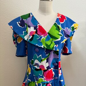 Op de afbeelding: Een blauwe jurk met een grote, kleurrijke bloemenprint en een brede kraag. De jurk heeft een dubbele knoopsluiting en korte mouwen.