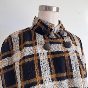 May include: A textured jacket featuring a plaid pattern in black, mustard yellow, and off-white. The garment has a high collar, a double-breasted front closure, and large, round, dark brown buttons arranged in a symmetrical grid pattern.