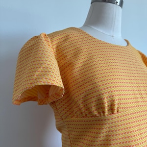 May include: A vintage-style dress in a vibrant yellow and orange pattern. The dress features short, ruffled sleeves and a fitted waist. The fabric has a textured appearance, and the dress is displayed on a mannequin.