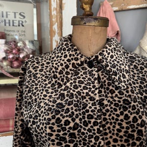 Puede incluir: Una chaqueta con estampado de leopardo en beige y negro sobre un maniquí vintage. La chaqueta tiene cuello y botones negros. El maniquí tiene una pieza dorada en la cabeza. El fondo incluye un letrero que dice "GIFTS FOR HER".