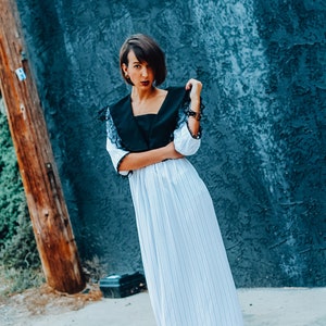 May include: A woman wearing a white and black striped maxi dress with a black lace collar. She is standing in front of a blue wall and wearing black platform sandals.