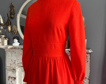 May include: A vibrant red vintage dress with a high neckline and long sleeves. The dress features a textured floral pattern and a fitted waist, with a flowing skirt. The dress is displayed on a mannequin.