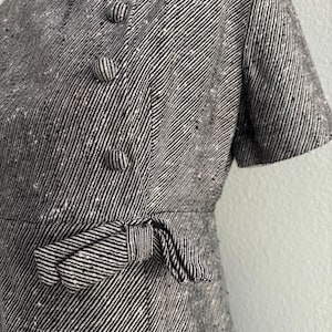 May include: A black and white tweed dress with short sleeves. The dress features a round neckline, decorative buttons, and a bow detail at the waist. The fabric has a textured appearance with diagonal stripes.