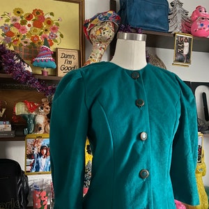 May include: A teal green blazer with a round neckline and long sleeves. The blazer has a button closure and features five buttons down the front. The blazer is on a mannequin.