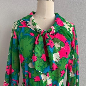 Puede incluir: Un vestido vintage con estampado floral en verde, rosa, azul y blanco. El vestido de manga larga presenta un cuello con lazo y un cinturón a juego. La tela tiene un patrón vibrante y colorido de flores y hojas grandes, creando un look retro audaz.