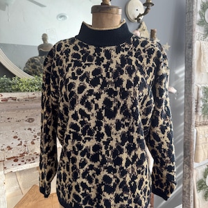 Puede incluir: Un suéter vintage de cuello alto con estampado de leopardo. El suéter presenta un cuello alto y puños negros, con un diseño de estampado animal en beige y negro. El suéter se exhibe en un maniquí.