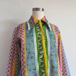 May include: A long-sleeve button-down shirt featuring a vibrant vertical striped pattern with alternating bands of floral motifs, paisley designs, and geometric diamond shapes in bright shades of blue, green, yellow, pink, and white.