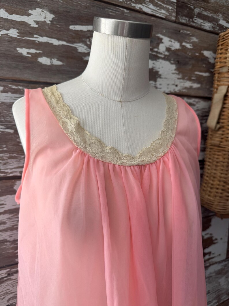 Op de afbeelding: Een vintage roze nachtjapon met een delicate kanten rand rond de halslijn. De mouwloze nachtjapon is gemaakt van een zachte, vloeiende stof en wordt getoond op een mannequin. De achtergrond omvat een houten muur en een rieten mand.