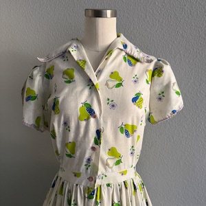 Op de afbeelding: Witte vintage jurk met een perenprint. De jurk heeft een kraag, korte mouwen en een geplooide rok. Het perenpatroon bevat groene, blauwe en gele peren met paarse bloemaccenten. De jurk is aan de voorkant dichtgeknoopt.
