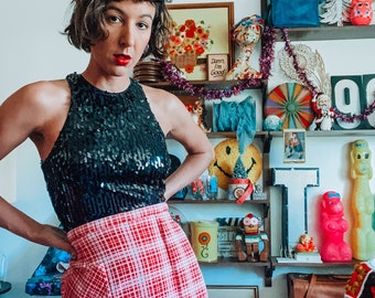 May include: A woman wearing a black sequined halter top and a red and white checked skirt. She is standing in front of a wall with a variety of decorations, including a framed picture, a smiley face, and a sign that says "Darn I'm Good."