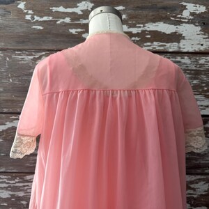Op de afbeelding: Een vintage roze nachtjapon op een mannequin. De doorschijnende stof heeft een hoge halslijn, korte mouwen met kanten afwerking en een vloeiend silhouet. De nachtjapon heeft een zachte, pastelroze kleur.
