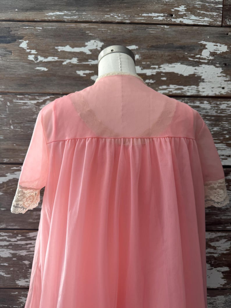 Op de afbeelding: Een vintage roze nachtjapon op een mannequin. De doorschijnende stof heeft een hoge halslijn, korte mouwen met kanten afwerking en een vloeiend silhouet. De nachtjapon heeft een zachte, pastelroze kleur.