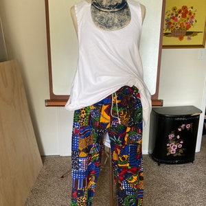 Op de afbeelding: Een mannequin toont een witte tanktop, een broek met patroon en een denim baseballcap. De broek heeft een levendig patchwork-ontwerp met blauwe, rode, gele en groene patronen. De pet is van verweerde blauwe denim. De outfit is casual en kleurrijk.