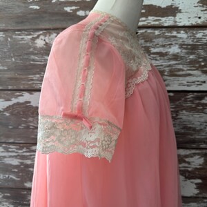 Op de afbeelding: Een vintage roze nachtjapon met korte mouwen. De nachtjapon heeft kanten afwerking rond de halslijn en manchetten. De mouwen zijn versierd met roze lintaccenten en kleine strikjes. Het kledingstuk wordt op een mannequin gepresenteerd.