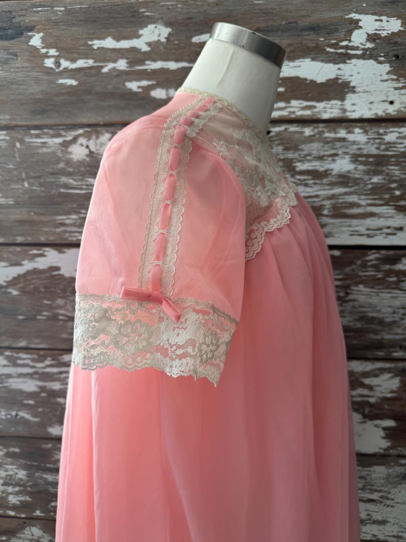Op de afbeelding: Een vintage roze nachtjapon met korte mouwen. De nachtjapon heeft kanten afwerking rond de halslijn en manchetten. De mouwen zijn versierd met roze lintaccenten en kleine strikjes. Het kledingstuk wordt op een mannequin gepresenteerd.