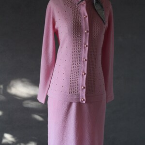 Peut inclure: Un ensemble jupe et cardigan en tricot rose avec un motif perl&eacute; et une fermeture &agrave; boutons. Le cardigan a des manches longues et un col rond. La jupe est mi-longue.