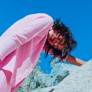 Peut inclure: Une femme portant un pull rose et un pantalon assorti pose devant une grande formation rocheuse. Le ciel est bleu azur.