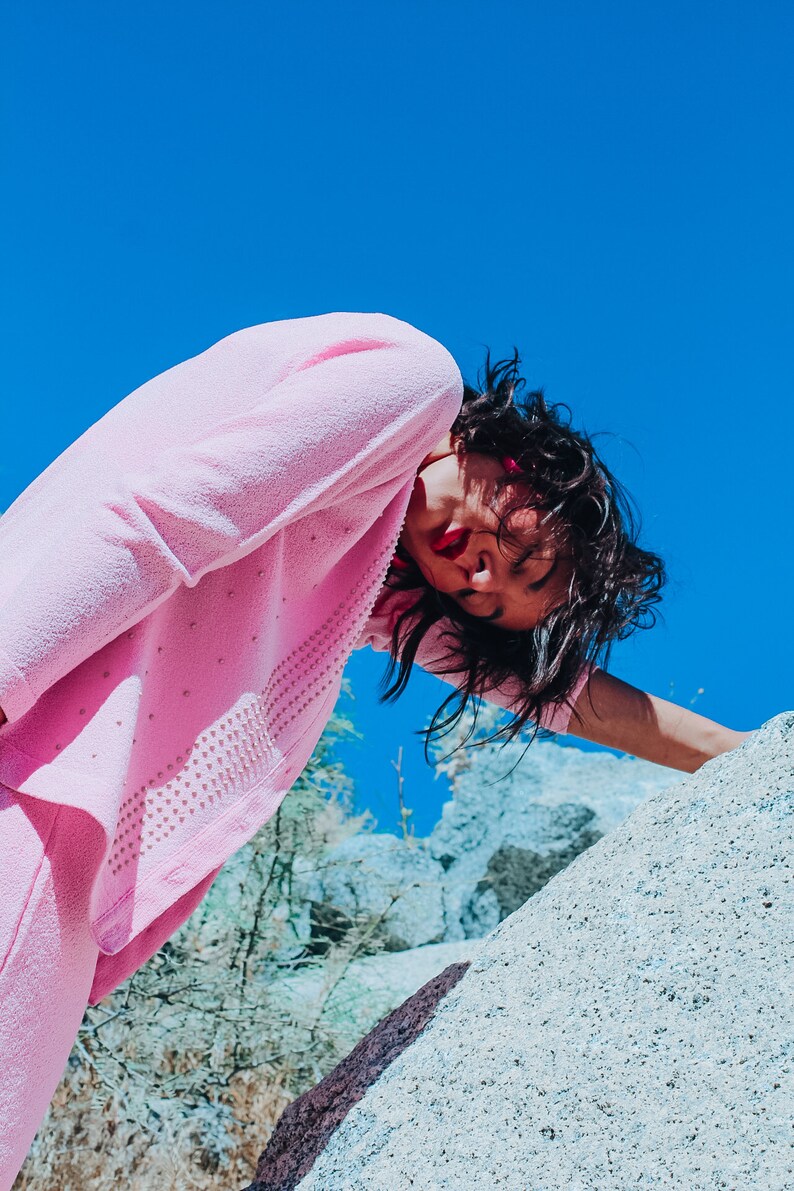 Peut inclure: Une femme portant un pull rose et un pantalon assorti pose devant une grande formation rocheuse. Le ciel est bleu azur.