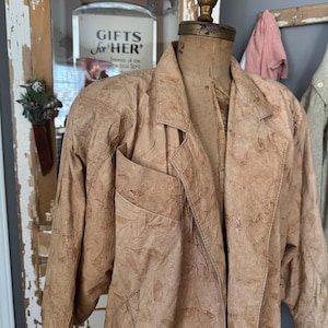 Puede incluir: Una chaqueta de color marrón claro, de estilo vintage, con un estampado de hojas, expuesta en un maniquí. La chaqueta presenta un cuello con solapas, un bolsillo en el pecho y un corte holgado. El maniquí tiene una cabeza de estilo vintage y está montado sobre un soporte de madera.