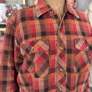 Puede incluir: Una camisa de franela a cuadros en tonos rojos, marrones y naranjas. Tiene cierre de botones y dos bolsillos en el pecho. La camisa se exhibe en un maniquí. Un estilo clásico, adecuado para uso informal.