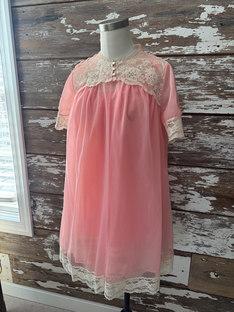 Op de afbeelding: Een vintage roze nachtjapon met korte mouwen en een kanten rand. De nachtjapon heeft een hoge halslijn met kanten details en kleine knopen. De zoom en manchetten zijn ook afgezet met kant. Het kledingstuk wordt op een mannequin getoond.