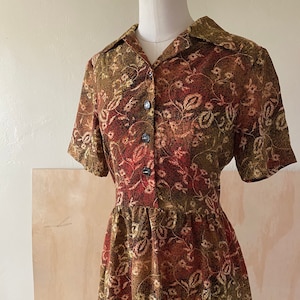 Puede incluir: Un vestido vintage de estampado floral con cuello, mangas cortas y cierre de botones en la parte delantera. El vestido está hecho de tela marrón y roja con un patrón de paisley.