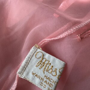 Op de afbeelding: Close-up van een doorschijnende, lichtroze stof met een aangehecht label. Het label is wit met goud geborduurde tekst met de tekst "Miss" en "WARN MACHINE DO NOT". De stof heeft een delicate, vloeiende uitstraling.