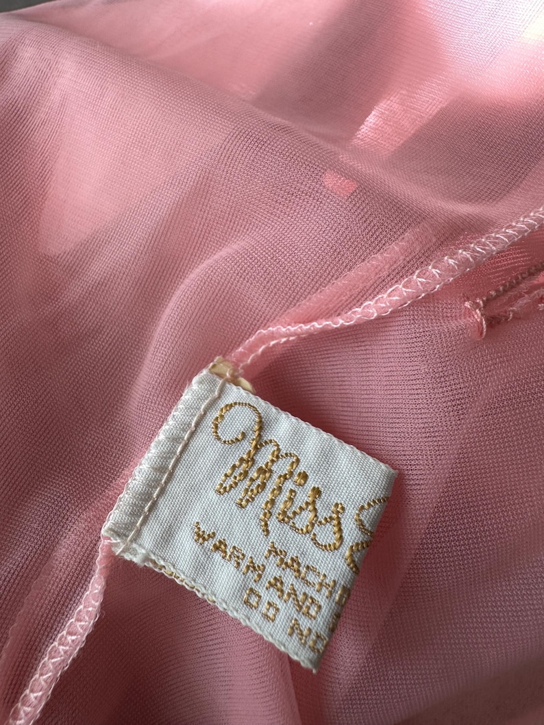 Op de afbeelding: Close-up van een doorschijnende, lichtroze stof met een aangehecht label. Het label is wit met goud geborduurde tekst met de tekst "Miss" en "WARN MACHINE DO NOT". De stof heeft een delicate, vloeiende uitstraling.