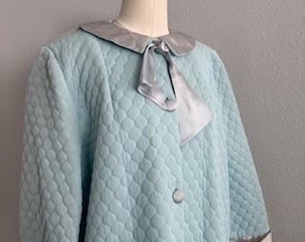 Op de afbeelding: Lichtblauwe gewatteerde ochtendjas met een zilveren satijnen kraag en bies. De ochtendjas heeft een strik bij de halslijn en drie knopen aan de voorkant. De mouwen zijn driekwart lang. De ochtendjas wordt op een mannequin getoond.