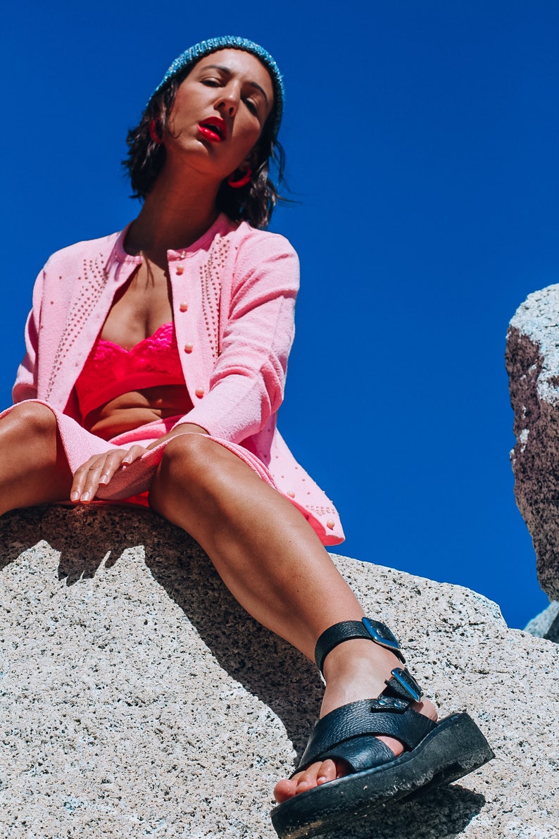 Peut inclure: Une femme portant un cardigan rose avec des perles, un soutien-gorge rose et des sandales &agrave; plateforme noires. Elle est assise sur un gros rocher avec un ciel bleu en arri&egrave;re-plan.
